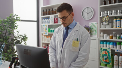 A young handsome hispanic man with a beard and glasses works behind a computer in a pharmacy,...