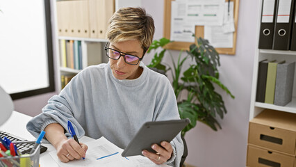 A focused hispanic woman with short blonde hair wearing glasses takes notes in a modern office.