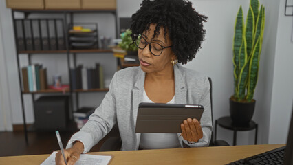 A young african american woman with curly hair in an office room, wearing glasses and a gray blazer, working intently on documents and a tablet at her desk.