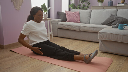A young african american woman with curly hair is sitting on a mat, exercising in a cozy living...