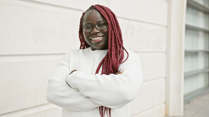 A smiling african woman with braids and glasses standing with arms crossed on an urban street.