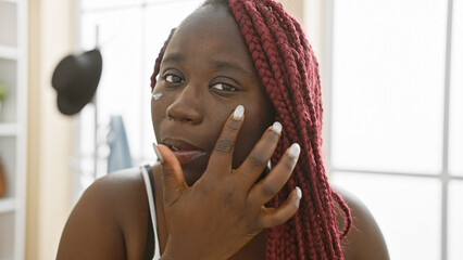An african american woman with braids applies makeup in a brightly lit room, reflecting beauty, femininity, and preparation.