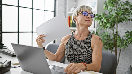 Mature woman cooling herself with paper in modern office setting, showing a sense of heat or frustration.