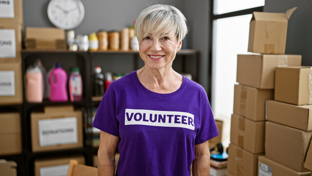 Mature woman with grey hair wearing volunteer shirt in a warehouse with boxes and donations.