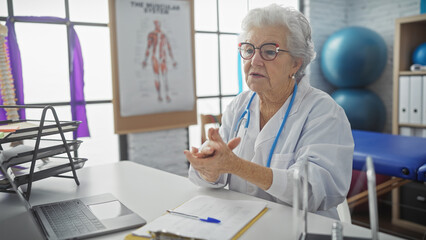 Senior woman doctor consulting in a modern clinic office with computer and medical poster.