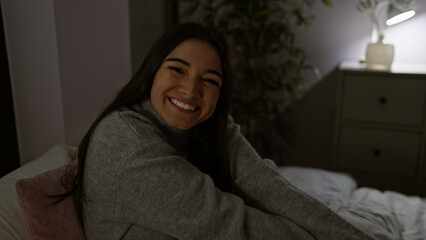 A smiling young hispanic woman sitting in a dimly lit indoor bedroom at night exuding warmth and...