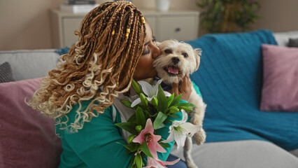 A young african american woman with braids lovingly kisses her small dog while sitting in a cozy indoor living room holding a bouquet of flowers.