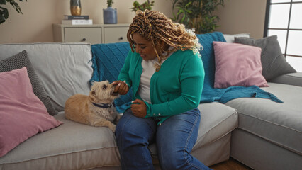 A young african american woman with braids sitting in a living room feeding her dog while they are on a comfortable sofa.