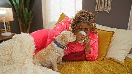 Fototapeta premium A young african american woman with braids relaxes on a bed in her bedroom, sharing a cup with her pet dog in a cozy indoor setting.