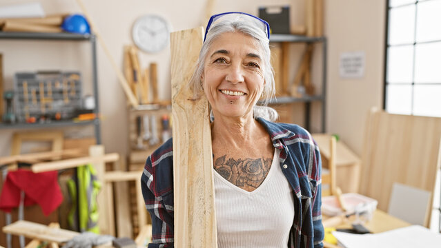 Smiling grey-haired middle age woman carpenter proudly holding a wood plank in her carpentry workshop