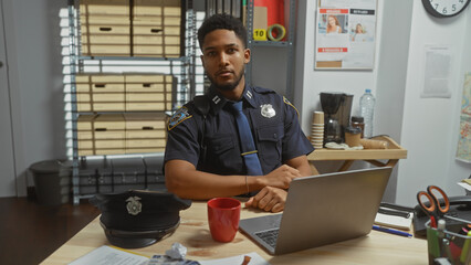 Handsome african american male police officer working on laptop in an office with evidence board.