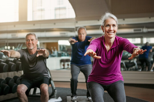 Mature group, woman and squat for exercise in gym, balance or coach for muscle development in class. Senior people, men and fitness for legs, smile or workout together in retirement at wellness club