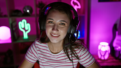 A smiling young woman with headphones in a colorful gaming room at night.
