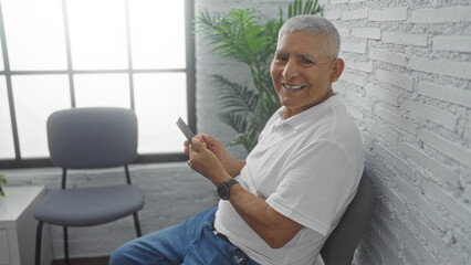 A handsome, grey-haired, hispanic man sits in a bright indoor room smiling and holding a phone, with a white brick wall and a window in the background.