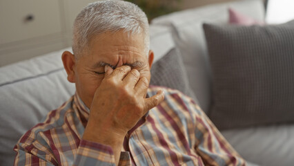A mature hispanic man with grey hair is rubbing his eyes while sitting on a couch in a cozy living...