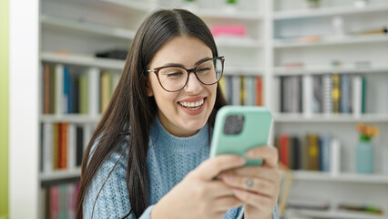 Young hispanic woman student smiling using smartphone at library university