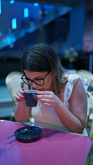 Charming hispanic woman in glasses enjoying her coffee break, sitting at a table in a bustling cafe at kabukicho, tokyo's iconic cityscape alive with skyscrapers