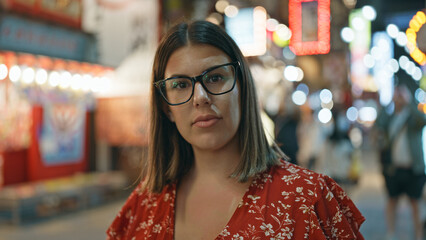 At night, on shinsekai street; a beautiful, serious hispanic woman with glasses stands, deep in...
