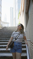 A smiling young woman in casual wear ascending stairs amidst dubai's modern skyscrapers exudes...