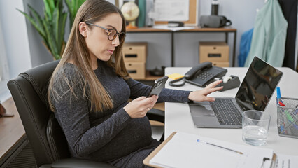 A pregnant hispanic woman examines her smartphone while working with a laptop in a modern office setting.