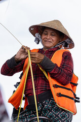 Vertical photo of a Vietnamese woman wearing an orange life jacket while making a bamboo leaf craft.