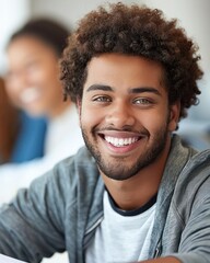 A young man with a bright smile and a beard sits in a classroom, looking directly at the camera
