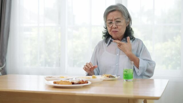 Handheld shot footage, An elderly Asian woman sat at the table eating fried food on a plate in front of her. She felt food stuck in her throat so she picked up her drink and drank it.