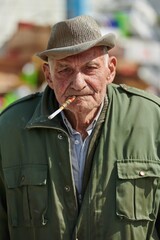 Obraz premium Older Man Smoking Cigarette in Dark Green Jacket and Cap