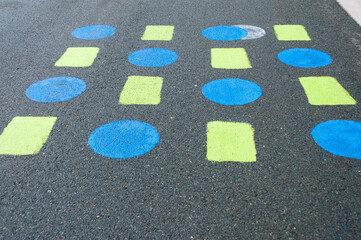 A black asphalt background with blue painted round markings and yellow squares. The children's educational game is on an outdoor playground surface. The activity improves kids' balance and jumping. 