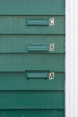 Three green metal mailboxes, postboxes, or letterboxes are affixed to a green apartment building. They are arranged in a row. The containers have the letters A, B, and B next to the slots.