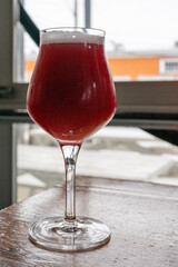 A red beer with a reddish or amber hue in a clear tulip glass with a long stem. The Irish Red Ale beer is aromatic and flavorful. The glass is on a wooden table with a colorful orange background.