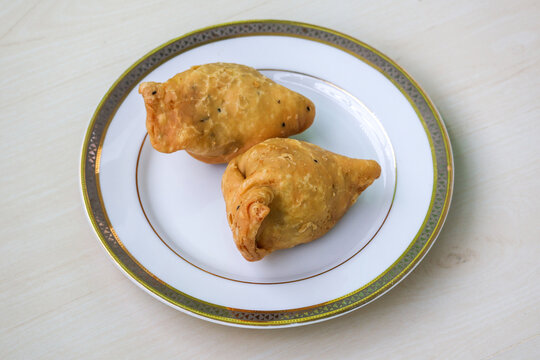Two fried samosas on a white plate on wooden surface. Traditional street food from Bangladesh and the Bengal region of India. It is also known as singara, singada, samose, somsa, sambusa, and samuza.