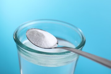 Spoon with baking soda over glass of water on light blue background, closeup