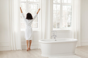 Morning routine. Black woman opening window in light cozy bathroom, lady wearing white bathrobe,...