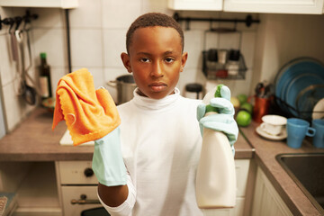 Angry upset black teen boy in white long-sleeve and blue rubber gloves holding rag and spray bottle...