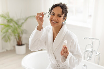 Latin lady applying moisturizing serum on face sitting at bathtub and making daily skincare...