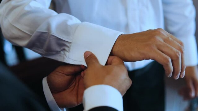 Close-up shot of unrecognizable best man putting silver cocoa bean-shaped cufflink in groom's tuxedo shirt 60fps