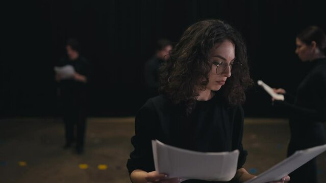 A confident girl with brown hair and round glasses in a black uniform suit reads her words and the plot of the play from sheets of paper during her preparation and rehearsal on stage in the theater