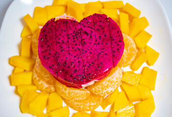 Red dragon fruit (pitaya) slices arranged in a heart shape, on top of mango and orange slices, on a white rectangular plate, on a wooden table. Celebrate Valentine in a healthy manner.