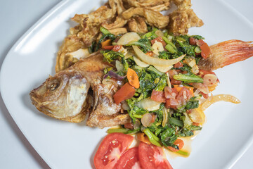 Indonesian food: fried nile tilapia (ikan nila goreng) with stir fried bok choy (sawi), crispy fried oyster mushroom (jamur tiram), and chopped tomatoes; on top of white rectangular plate
