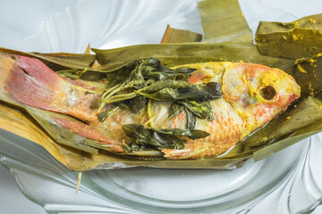 Indonesian food: pepes ikan nila (steamed nile tilapia with spices). Spices are smeared on tilapia's body, wrapped with banana leave, and then steamed. On top of a glass plate and white tablecloth.