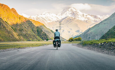 Back view one male cyclist in blue jacket pedaling on touring bicycle surrounded in mountains and green summer nature. Bicycle tour touring cycling holidays