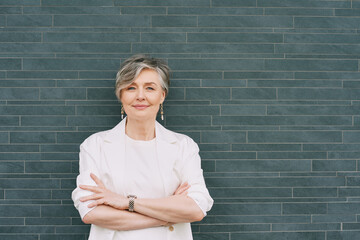 Portrait of 55 - 60 year old woman with grey hair, wearing white jacket, posing on grey background