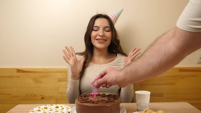 The girl is sitting in front of a table with a festive cake, in which a candle in the form of the number 19 is burning, which she blows out. Birthday celebration concept. Slow motion