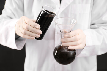 Woman pouring black crude oil from beaker into flask on dark background, closeup