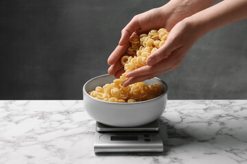 Woman adding pasta into bowl on kitchen scale on white marble table, closeup