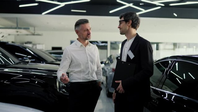 A confident guy a brunette assistant in glasses and a black body suit, communicates with a middle-aged man in a white shirt about a specific model of a modern white car in a car showroom