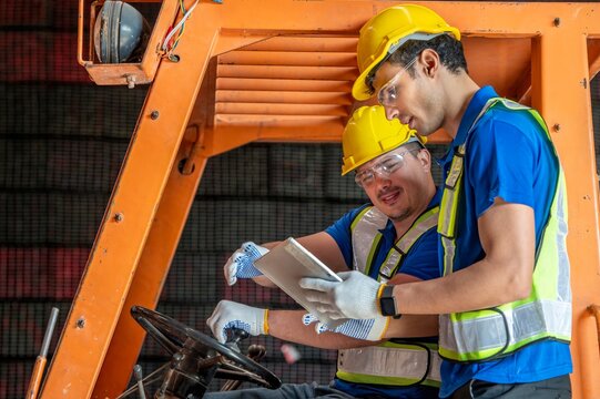 Two warehouse workers wearing hard hats and safety vests are using a tablet computer to check inventory while sitting on a forklift.