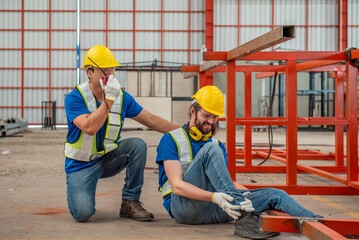 A worker is injured and sits on the ground holding his leg while another worker kneels beside him...