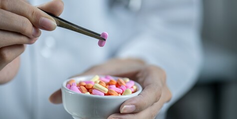 A close-up image of a doctor holding a pink pill with tweezers over a white bowl filled with pills.